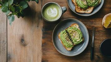 Top-down shot of two avocado toasts on gray plates, a matcha latte, and a knife on a textured wooden table. Healthy brunch setting with plant photo