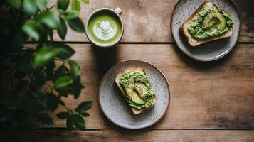 Overhead view of two avocado toasts on speckled plates with a matcha latte on a rustic wooden table. Healthy and vibrant breakfast scene photo