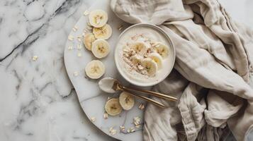 Creamy banana yogurt bowl with oatmeal, cinnamon, and fresh banana slices on marble table with fabric and golden cutlery photo