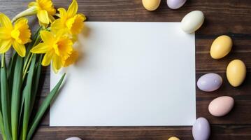 Yellow daffodils and pastel Easter eggs placed around blank white paper on rustic wood background, symbolizing spring celebration and greeting Space for text photo