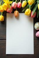 Colorful tulips and painted Easter eggs surround empty white sheet on dark wood table, perfect for spring or holiday-themed message or design Space for text photo