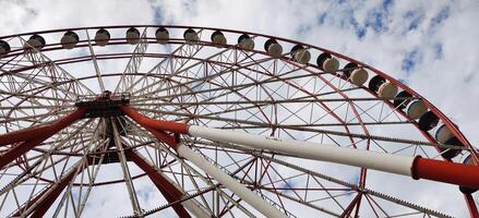 Low-angle view of a large Ferris wheel with red and white metal beams against a partly cloudy sky, showcasing its towering structure and passenger cabins. photo