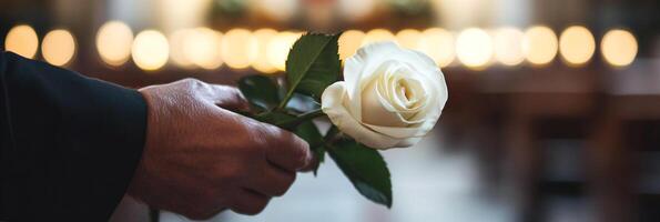 Hand of a man in a black suit holding a white rose, with a church in the background. Concept of a groom awaiting his bride, wedding atmosphere. photo