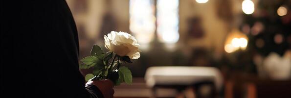 Man wearing a black suit and holding a white rose stands in a church setting. Banner concept representing mourning, honoring a loved one, and the farewell at a funeral. photo