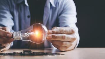New idea concept with coins stack save power to save the earth, young Asian men hand holding light bulb with new knowledge light bulb and money stack on wood table on black background with copy space photo