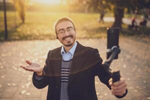 Male influencer explaining his business success through blogging, smiling and gesturing while recording with his smart phone and gimbal in a park. photo