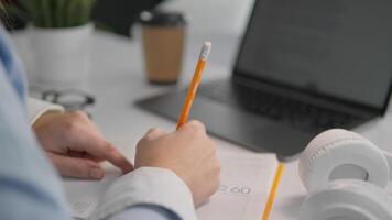 In a focused study environment, a dedicated individual is taking notes with a pencil on paper. A laptop and coffee cup nearby enhance the productive atmosphere and engaged learning experience video