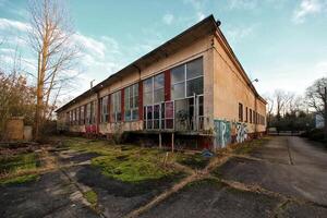 Abandoned glass building with graffiti-covered walls, overgrown pavement, and broken windows photo