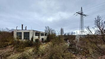 Deteriorating industrial site with overgrown vegetation and a power line tower in the background photo