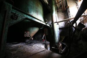 Rusted green tank beneath corroded pipes in a dimly lit corner of an abandoned factory photo
