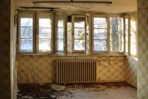 Broken old windows in a decayed room with peeling wallpaper and a rusted radiator below photo