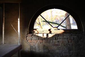 Arched brick window letting in sunlight through a broken frame with an exposed weathered wall photo