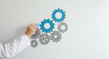 Hand in lab coat draws connecting blue and grey gears with chalk on a whiteboard, representing teamwork, process, and system building. photo