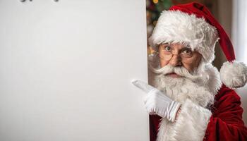 Santa Claus in red suit and hat points to a large blank white list or sign with copy space for text or message. photo