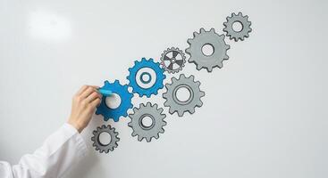 Hand in lab coat draws blue gear connecting to grey gears on whiteboard, symbolizing system and teamwork. photo