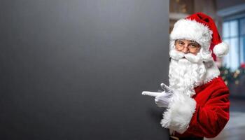 Traditional Santa Claus peeking from behind a large dark gray vertical surface, pointing left with copy space for text or image. photo