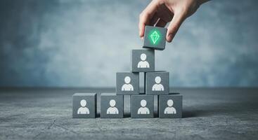 Hand placing a block with a diamond icon atop a pyramid of blocks with businessman icons, representing talent, leadership, and hierarchy. photo