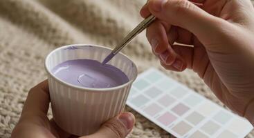 Close up on a hand holding a paint brush dipping into a cup of purple paint, with a color palette nearby, showcasing color selection. photo