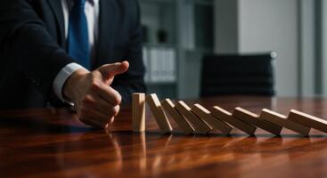 Businessman stopping wooden dominoes from falling on a desk, symbolizing crisis management, risk prevention, and business continuity planning. photo
