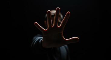 Man raises hand in front of face in dark setting, fingers spread. Gesture of stopping, blocking, or Vulcan salute concept on black background. photo
