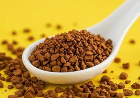 Close up of instant coffee granules on a white ceramic spoon against a bright yellow backdrop. Coffee granules are also scattered around the spoon. photo