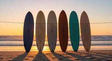 Six surfboards stand in a row on a sandy beach at sunset, casting long shadows. Ocean waves roll in the background under a colorful sky. photo