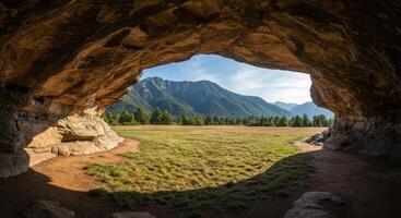 Cave entrance overlooking a grassy field with mountains and trees in the distance under a blue sky. Landscape with natural rock formations and scenic view. photo