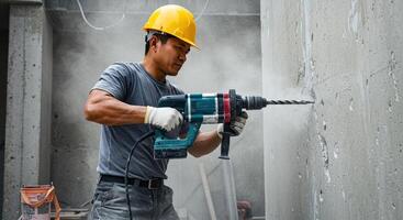 Construction worker drills into concrete wall on construction site. He is wearing a yellow hard hat and gloves for safety. Dust is flying as he works. photo