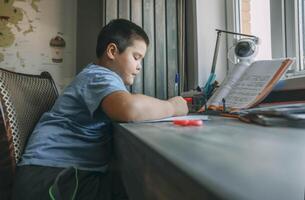 asian boy write in notebook, homework beside open textbook on desk, study tool, education, homeschooling, parenting resource photo