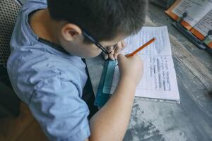 boy in blue shirt using ruler and pencil while doing homework, close up of child writing in notebook, wooden desk with textbook photo