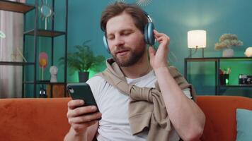 Happy caucasian man in wireless headphones relaxing sitting on sofa at home listening favorite music video