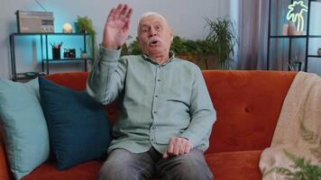 Grandfather man smiling friendly at camera, waving hands gesturing hello hi greeting at home on sofa video