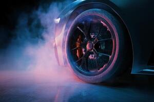 Close-up of a car wheel with alloy rim, illuminated by vibrant blue and pink lights. Smoke surrounds the tire on a reflective floor, creating a dynamic and futuristic atmosphere photo