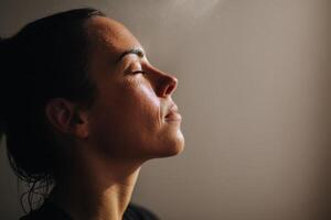 A woman with closed eyes and serene expression with soft lighting highlighting her profile and skin texture. Her hair is tied back, and she appears in a moment of calm reflection photo