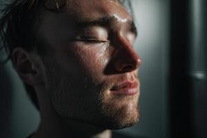 Wet face with closed eyes in solemn expression, highlighted by dramatic lighting, showing textured skin and light beard. The image conveys a sense of serenity and introspection photo