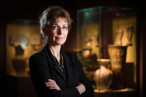 Curly-haired woman with glasses and a confident expression, wearing a black outfit with arms crossed, stands in front of display cases with ancient artifacts and pottery in museum photo