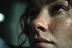 Face of a person with a serious expression, focused on the eyes and skin texture. Curly hair partially visible, with a shadowy background suggesting an indoor setting photo