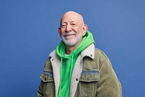 Smiling bald man with a gray beard wears a green hoodie and a jacket with shearling collar. His expression exudes warmth and friendliness against a simple setting photo