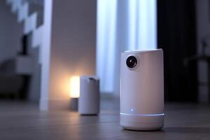 A small cylindrical security camera with a visible lens sits on a wooden floor indoors. Another similar device glows softly in the background, both near a staircase photo
