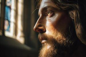 Detailed close-up of a statue's face with strong natural light highlighting its features, showcasing a bearded figure gazing solemnly. A blurred stained glass window appears in the background photo