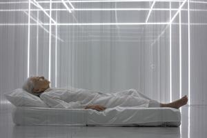Elderly woman lies on a white mattress in a minimalistic, futuristic room with glowing line structures. She wears white pajamas and rests with closed eyes, creating a tranquil atmosphere photo