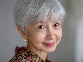 Smiling elderly woman with short gray hair and delicate earrings is wearing a patterned blouse. She has bright eyes and a gentle expression, with a soft focus background. No text visible photo