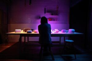 Silhouette of a person sitting at a table with various objects under soft, colorful lighting. Shelves and kitchen items are visible on the wall. The room is dimly illuminated photo