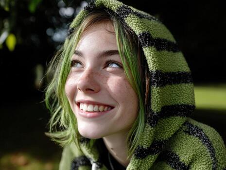 Smiling person with green hair and freckles looks up while wearing a striped green hoodie. Sunlight highlights the details, adding contrast between the subject and the dark background photo
