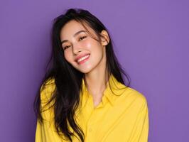 Smiling woman with long black hair wears a vibrant yellow shirt, head tilted slightly, against a solid backdrop. Her expression is cheerful and relaxed, with natural makeup highlighting her features photo