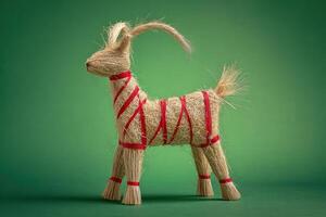 Straw goat figure wrapped with red ribbons stands against a green surface. The handcrafted piece displays intricate texture and detail, featuring distinctive long tail and upright posture photo
