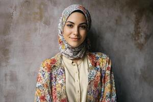 Smiling woman wearing a floral-patterned hijab and matching jacket over a pale shirt, standing against a textured wall, exuding elegance and confidence with a peaceful demeanor photo