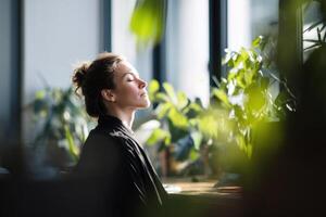Peaceful woman with closed eyes, sitting indoors surrounded by lush green plants with sunlight filtering through windows. She has a calm expression, and hair is tied up neatly photo