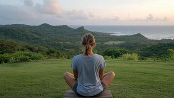 A woman sitting in a meditation pose on a grassy hill overlooking the ocean photo