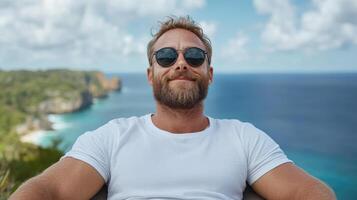 A man with sunglasses and a beard sitting on a chair with the ocean in the background photo
